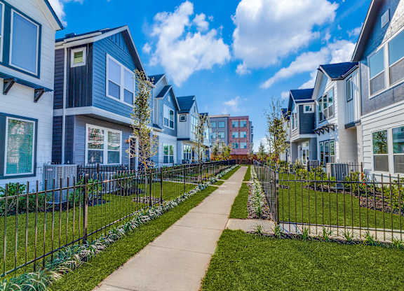 Houses for Rent in Houston, TX - East Heights - Exterior View of Apartment Homes with Fences, Manicured Grass Yards, Concrete Walking Path, and Trees.