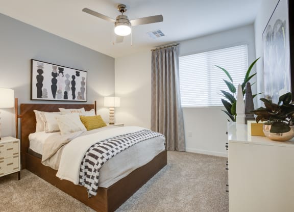 One-Bedroom Apartments in South Scottsdale, AZ - Lucent Scottsdale - Spacious Bedroom With Carpeted Flooring, Bed, Nightstands With Lamps, White Dresser, Ceiling Fan, And Window With Blinds.