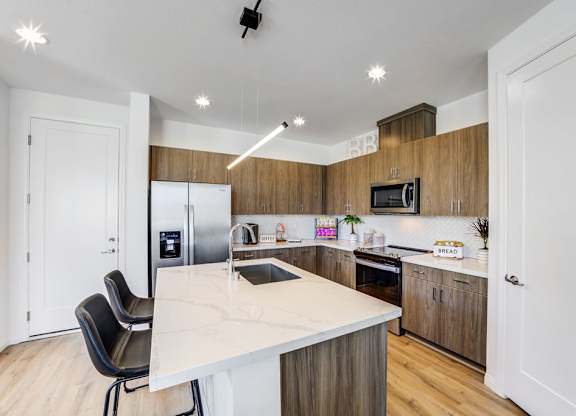 New Luxury Apartments In Skye Canyon, NV - Banyan Brighton - Open-Concept Kitchen With Wood-Inspired Flooring, Marble Countertops, Hardwood Cabinets, Tile Backsplash, Energy Efficient Appliances, A Kitchen Island, And A Pantry.