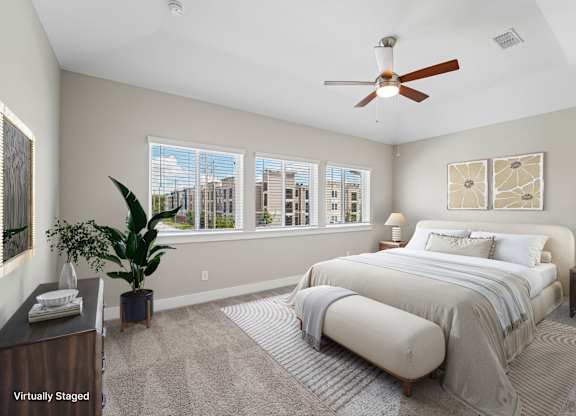 A bedroom with a large bed, a ceiling fan, and a view of the city through the windows.