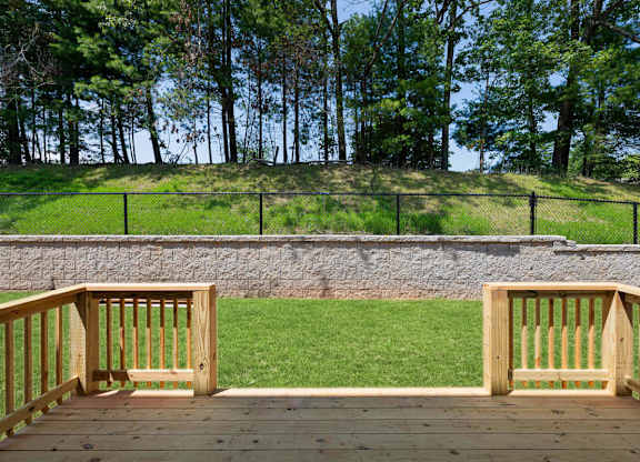 A wooden deck overlooks a grassy area with a stone wall and trees in the background.