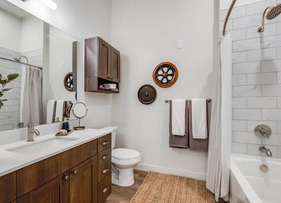 1 Bedroom Luxury Apartments in Greeley, CO - The Shelby - Bathroom with Wood-Style Flooring, High Ceiling, White Quartz Countertop Sink with Brown Wood-Style Drawers and Cabinetry, Large Mirror, Wall-Mounted Cabinets, and White Tile-Lined Shower with Tub and Shower Curtain.