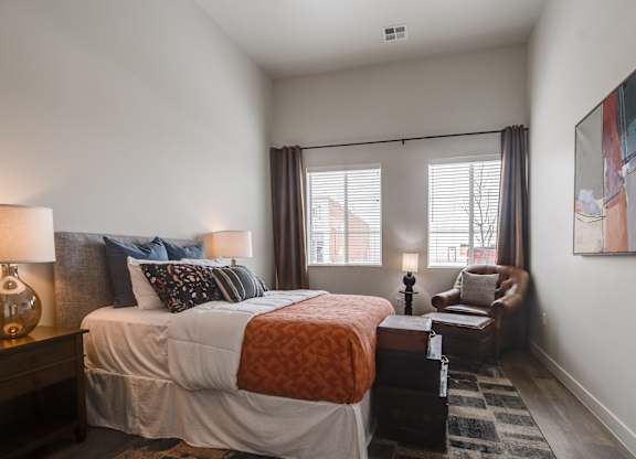 One Bedroom Luxury Apartments in Greeley CO - The Shelby - Furnished Bedroom with a Large Bed, a Leather Chair, Two Windows, White Walls, a Wooden Nightstand, and Wood-Style Flooring