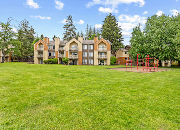 A playground with a red swing set is in the foreground of a grassy area in front of apartment buildings at MonteVista Apartments, Beaverton, OR 97007
