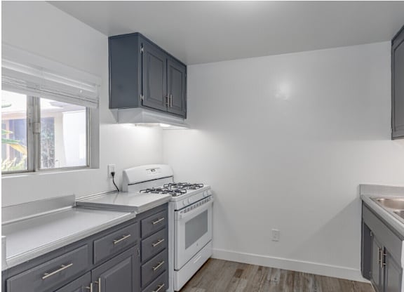 El Cajon, CA Apartments - Ivanhoe Village Apartments - Compact Kitchen With Gray Cabinetry, White Gas Range, Double Sink, Light Countertops, And Wood Style Flooring.