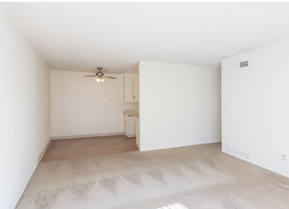 One-Bedroom Apartments in El Cajon, CA - Ivanhoe Village Apartments - Bright Bedroom With Neutral Carpet, White Walls, Ceiling Fan, And Adjacent Kitchen Entry.
