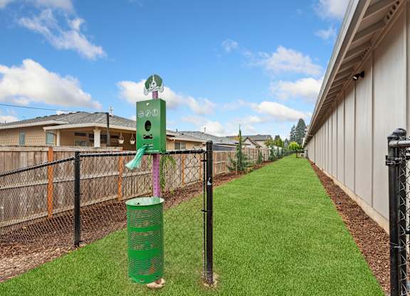 A green trash can sits on a grassy area next to a fence.