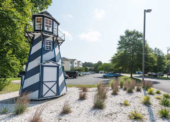 A blue and white striped lighthouse sits on a gravel area in front of a street.