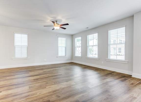 Single Family Homes in North Charleston, SC - Sumner Village - Empty Living Room Area with Hardwood-Style Flooring, a Ceiling Fan with Lighting, and Multiple Large Windows with White Blinds.