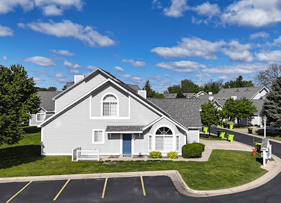 A house with a grey roof and a blue door is surrounded by a parking lot.