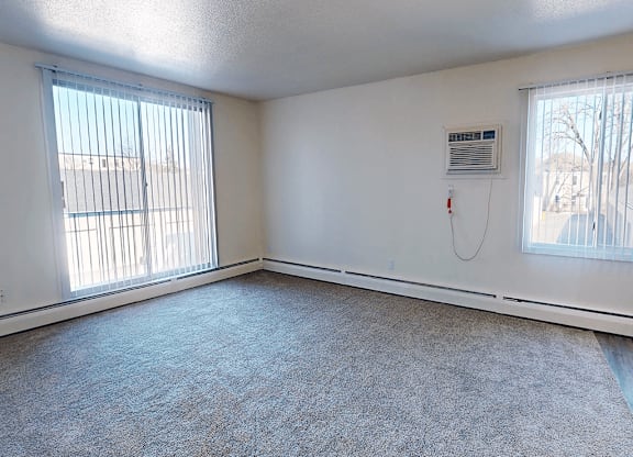 A room with a carpeted floor, two windows with blinds, and a wall-mounted air conditioning unit.