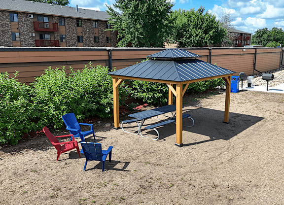 A gazebo with a black roof is surrounded by four chairs and a bench in a sandy area.