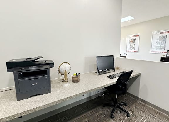 A black printer sits on a desk next to a black laptop and a black chair.