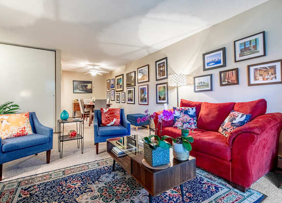 A living room with a red couch, blue chairs, and pictures on the wall.