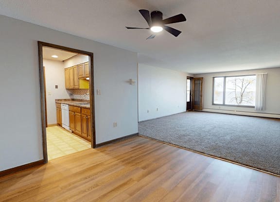 A room with a ceiling fan and a kitchen area visible through the door.