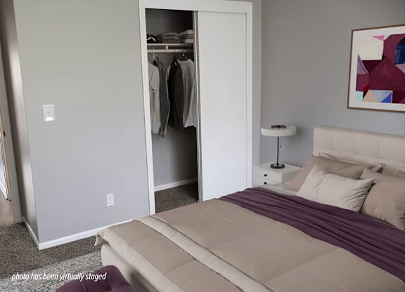 A bedroom with a bed, nightstand, lamp, and a closet with clothes hanging.