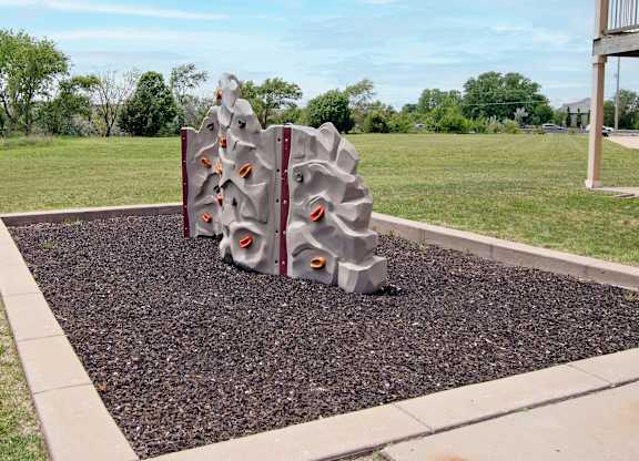 A playground structure with a grey and red color scheme is situated in a gravel area with a grassy background.