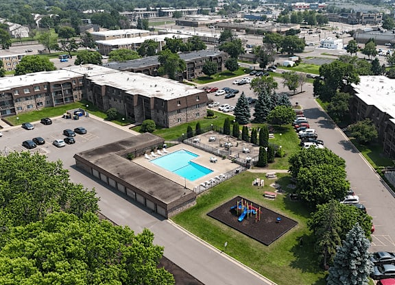 A swimming pool is surrounded by a playground and trees in a parking lot.