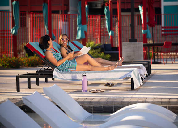 Two women are sunbathing and reading a book by the pool.