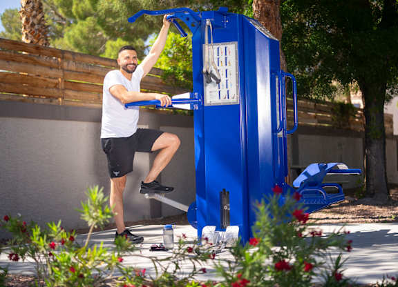 A man in a white shirt and black shorts is exercising on a blue gym machine outdoors. at The 95 Apartments, Las Vegas