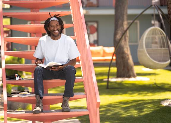 A man with dreadlocks is sitting on a red staircase reading a book.