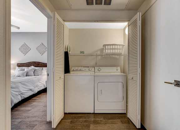 A white laundry machine is in a small room with a bed in the background.