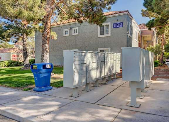 A blue trash can is in front of a grey building with a tree in front of it.