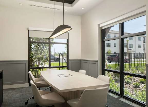 A conference room with a long table and chairs, a hanging light fixture, and a view of a parking lot through a glass door.