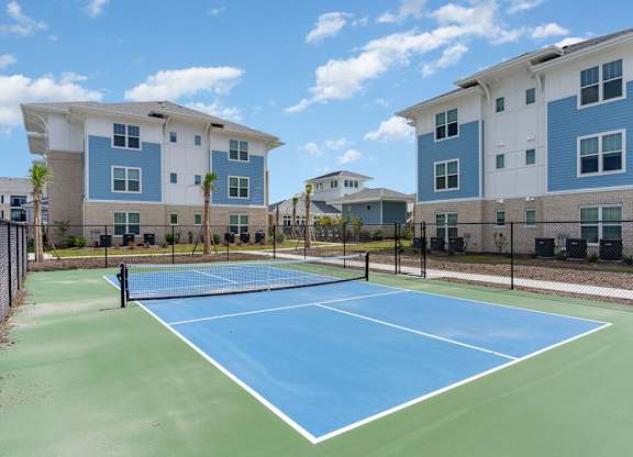 A tennis court is surrounded by two identical buildings.