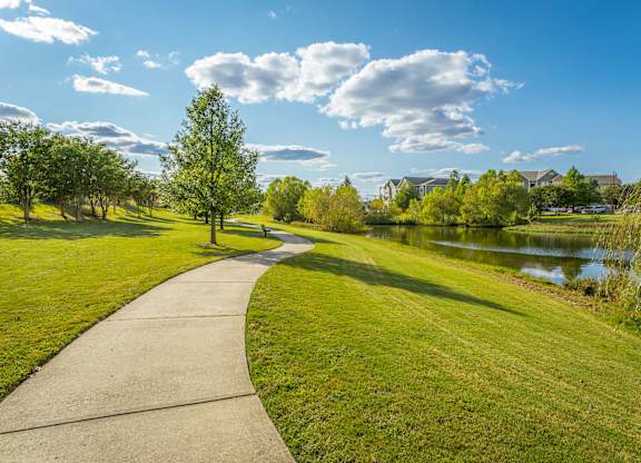 A concrete walkway leads through a grassy park with a lake and trees in the background.