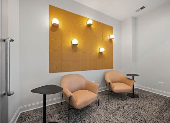 Two tan chairs and a small table are in front of a yellow wall with four lights on it.