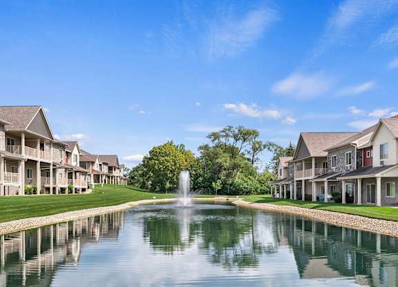 A serene scene of a pond with a fountain in the middle of a row of houses.
