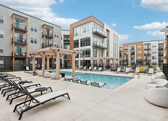 A pool area with sun loungers and a pergola in front of apartment buildings.