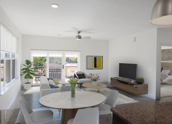A dining room table surrounded by four chairs, next to a furnished living room with a ceiling fan and a sliding glass door.