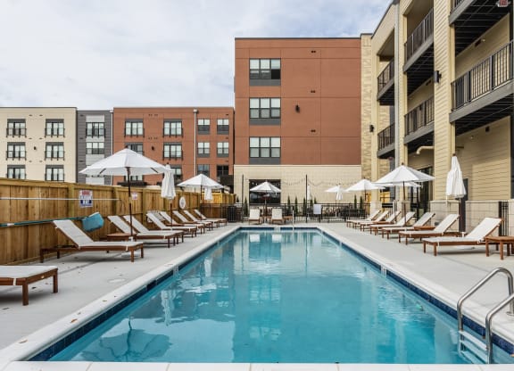 A swimming pool surrounded by a fence, white chairs and white umbrellas, next to apartment buildings.