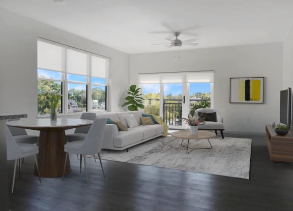 A spacious living room with a white couch and chair, a rug on hardwood-style floors, and large windows, next to a dining room table.