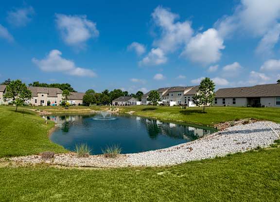 A pond in the middle of a grassy area with houses in the background.