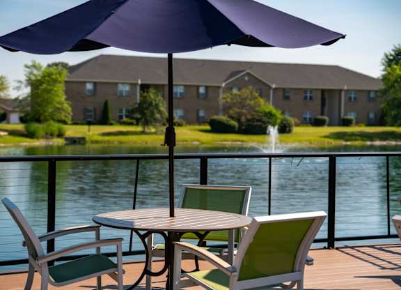A table with a purple umbrella is set up on a deck overlooking a pond.