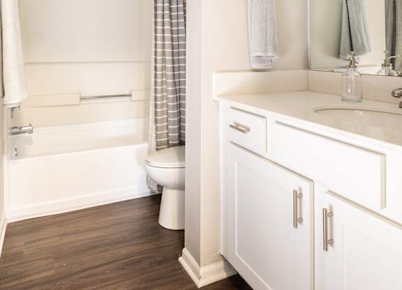A bathroom equipped with a white vanity with a mirror, a white tub, and hardwood flooring.