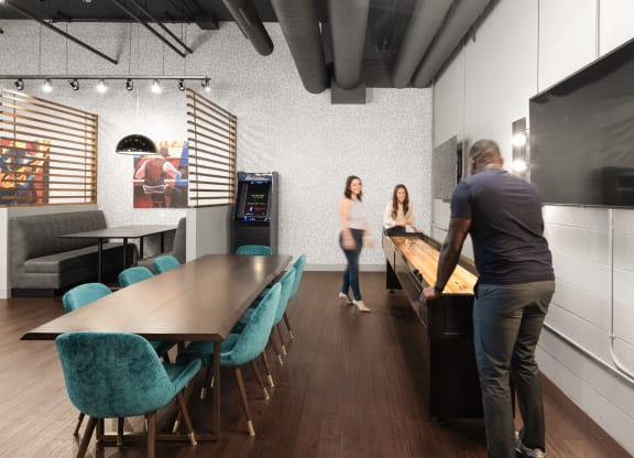A community lounge with a game center and a table surrounded by eight blue chairs on hardwood-style flooring.