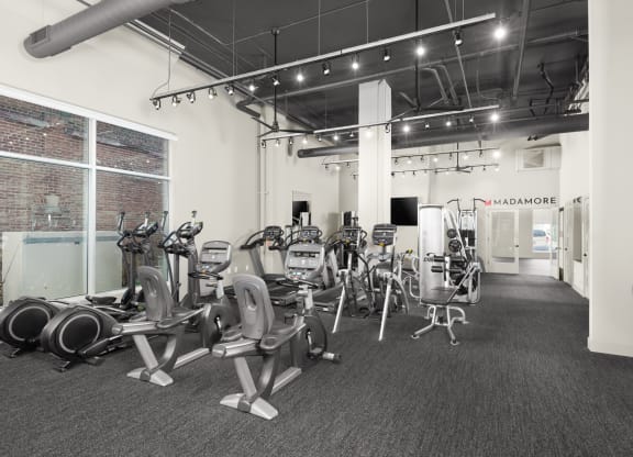 A fitness center equipped with a selection of workout machines on carpeted flooring, and large windows.