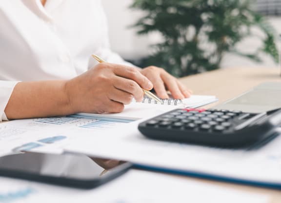 A person in a white shirt is using a calculator at a desk with papers and a laptop.