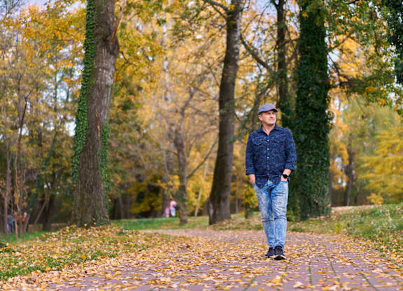 A man in a blue shirt and jeans stands on a path in a park with trees and ivy.