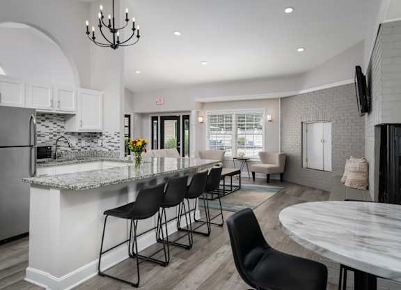 A kitchen in a clubhouse with stainless steel appliances and four barstools, next to a seating area with two chairs.