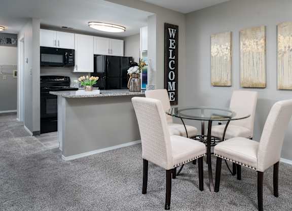 Dining room circular table surrounded by four chairs, situated next to a kitchen, on carpeted flooring.