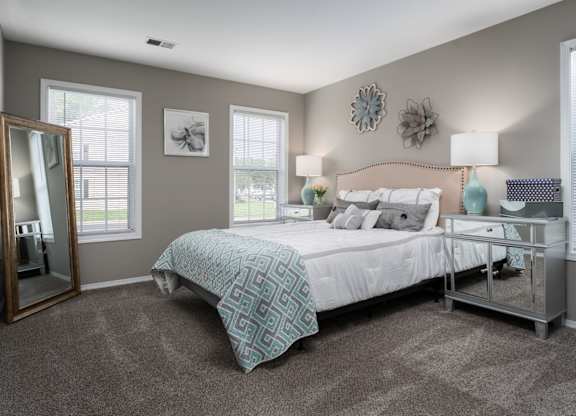 A bedroom with a bed and two bedside tables with a lamp on each side, carpeted floors, and two windows.