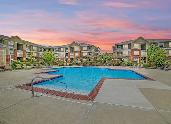 Swimming pool surrounded by chairs, next to apartment buildings, and a sunset.