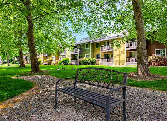 a park bench on a gravel path in front of a yellow apartment building