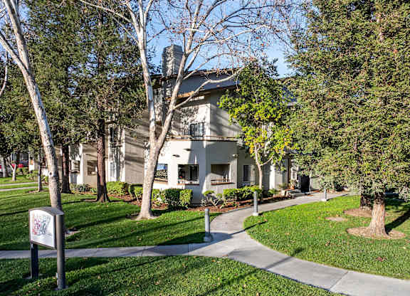 A tree-lined street with a white house and a sign on the sidewalk at The Woods Apartments, San Jose, 95136
