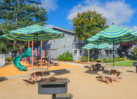 A playground with a green slide and two blue and white striped umbrellas at Cherrywood Apartments, San Jose, 95118